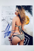 Signed full photo of Hailey Clauson posing against a graffiti-marked brick wall wearing a patterned bikini and beaded bracelets, featuring a blue autograph.