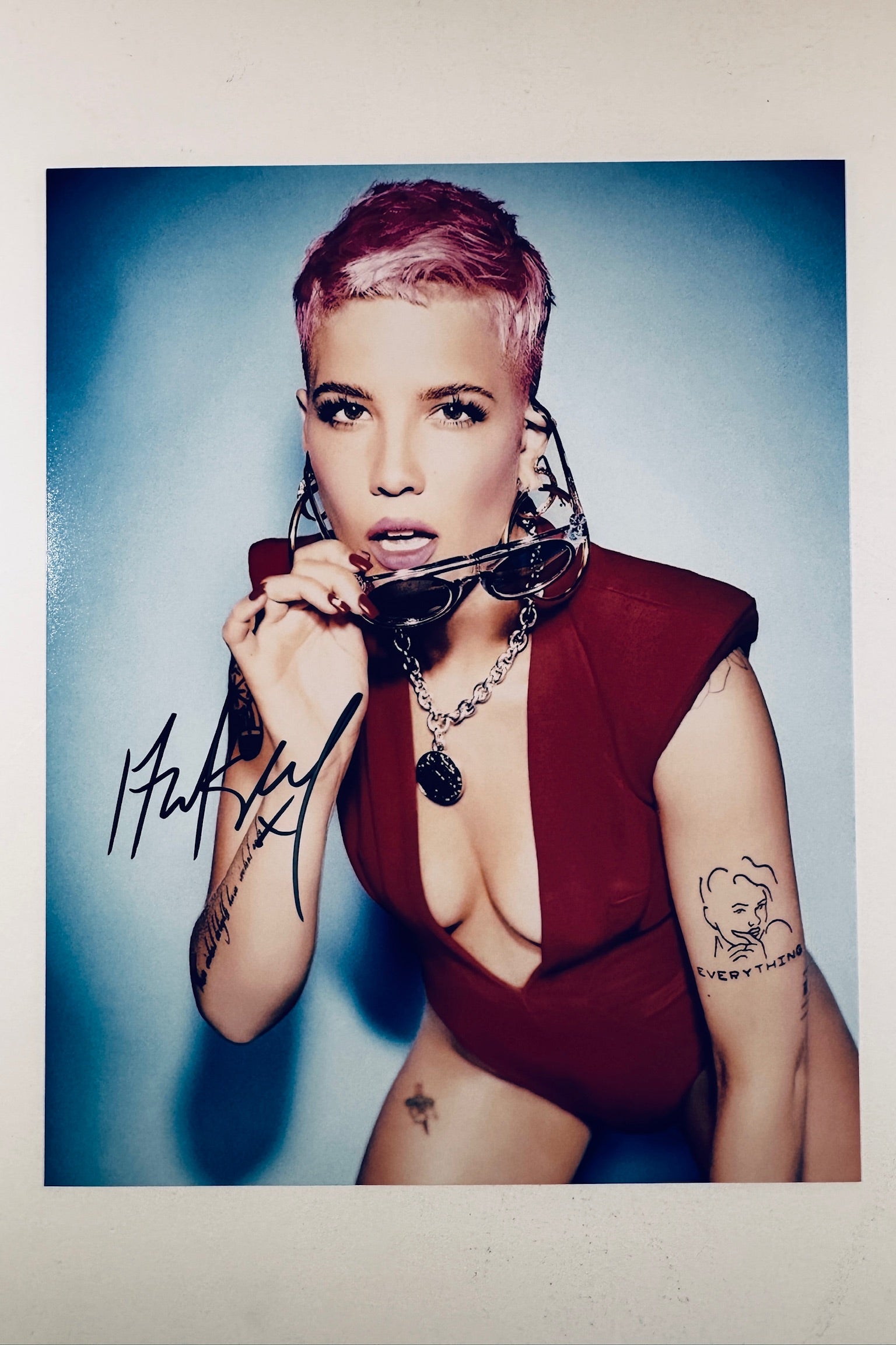 A portrait of Halsey with short pink hair, wearing a dark red revealing top and holding sunglasses, featuring a black signature on the lower left.