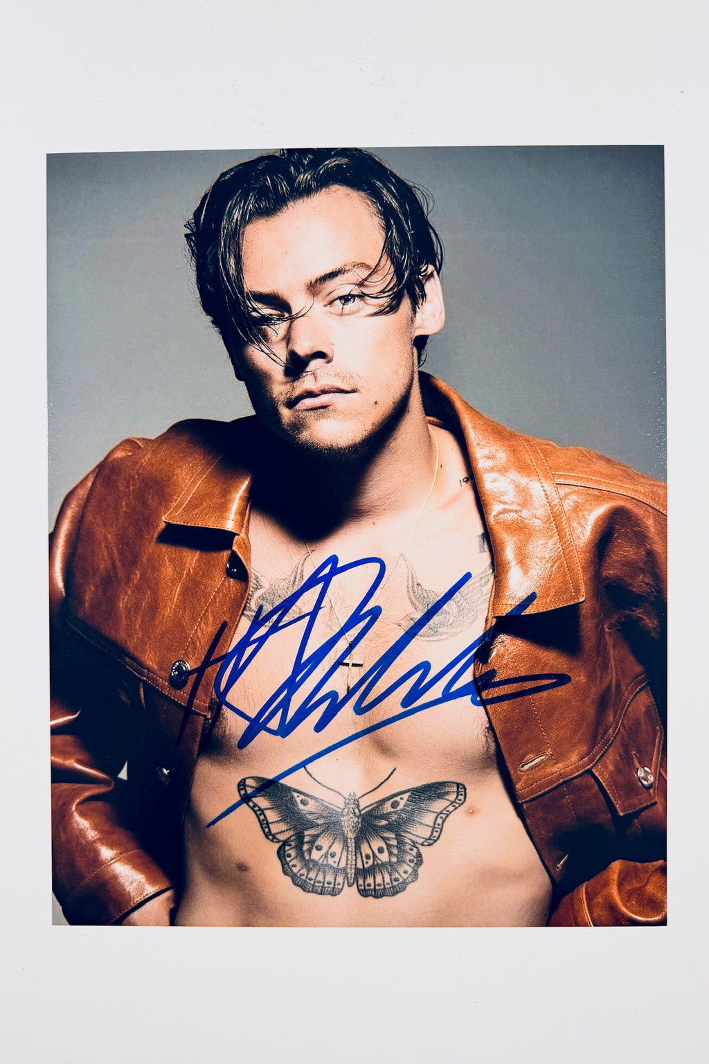A portrait of Harry Styles wearing an unbuttoned brown leather jacket with his butterfly chest tattoo visible, featuring a blue marker signature across his chest and torso.