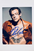 A portrait of Harry Styles wearing an unbuttoned brown leather jacket with his butterfly chest tattoo visible, featuring a blue marker signature across his chest and torso.