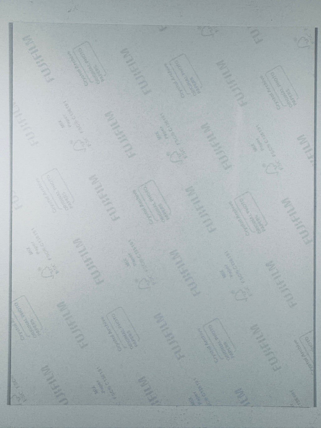 A light gray Fujifilm protective film or cover sheet with repeating diagonal brand logos and small printed icons, framed by thin dark vertical lines on the left and right edges.
