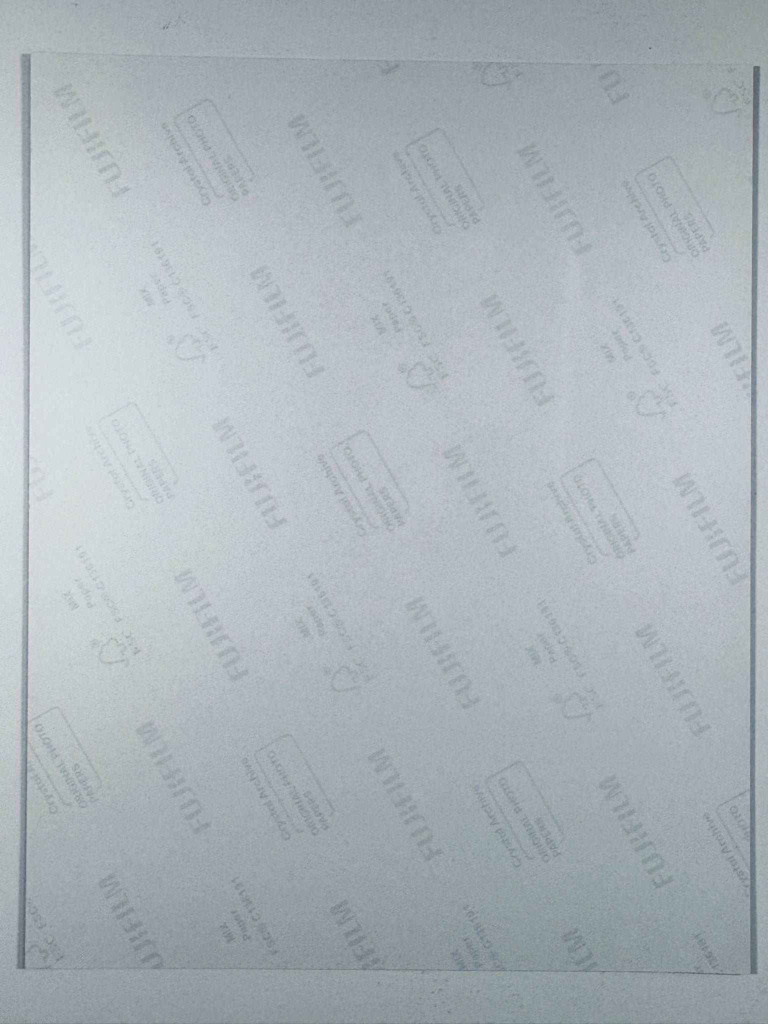 A light gray Fujifilm protective film or cover sheet with repeating diagonal brand logos and small printed icons, framed by thin dark vertical lines on the left and right edges.