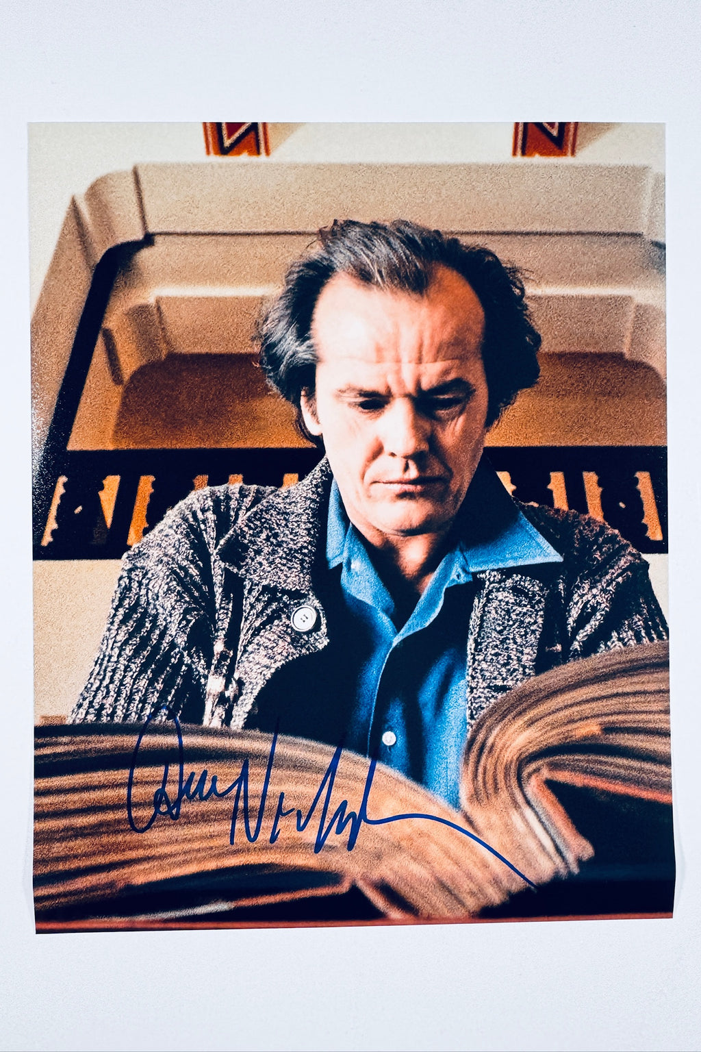 A still from The Shining featuring Jack Nicholson as Jack Torrance reading a large book with a tense expression, signed in blue ink across the bottom of the open book.