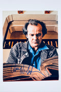 A still from The Shining featuring Jack Nicholson as Jack Torrance reading a large book with a tense expression, signed in blue ink across the bottom of the open book.