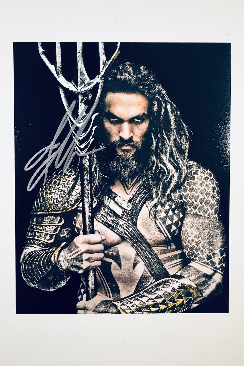 A portrait of Jason Momoa as Aquaman with long hair and a beard, wearing armor and holding a trident against a dark background, featuring a silver marker signature across the upper left and trident handle.
