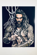 A portrait of Jason Momoa as Aquaman with long hair and a beard, wearing armor and holding a trident against a dark background, featuring a silver marker signature across the upper left and trident handle.