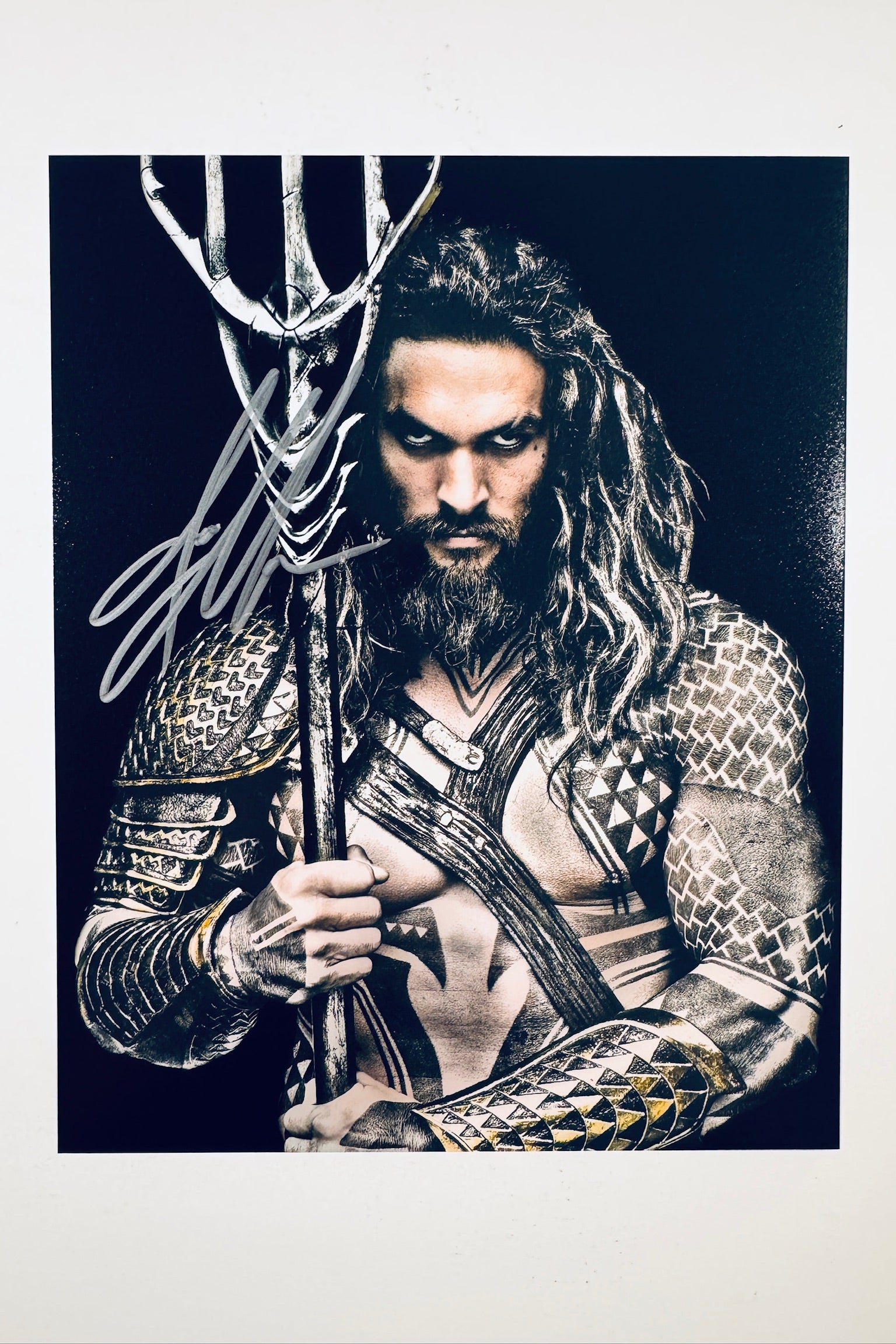 A portrait of Jason Momoa as Aquaman with long hair and a beard, wearing armor and holding a trident against a dark background, featuring a silver marker signature across the upper left and trident handle.