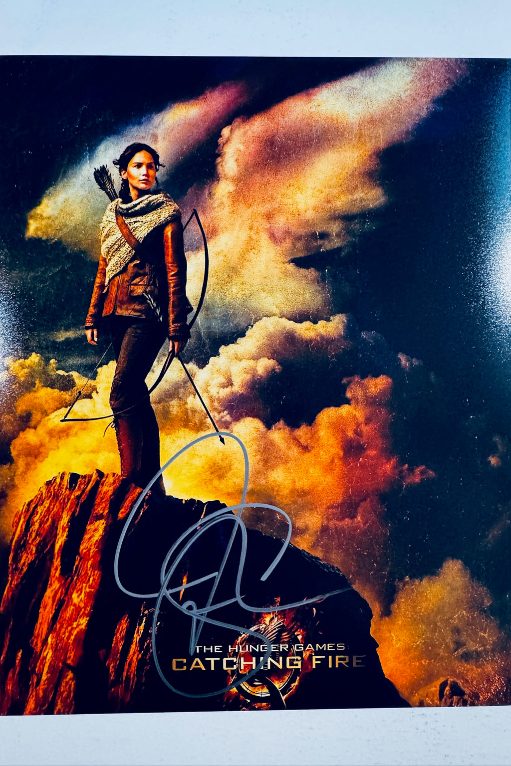 A portrait of Jennifer Lawrence as Katniss Everdeen from The Hunger Games: Catching Fire, standing on a rocky outcrop holding a bow and arrow against a dramatic sunset sky with fiery clouds, featuring a dark marker signature across the lower center.