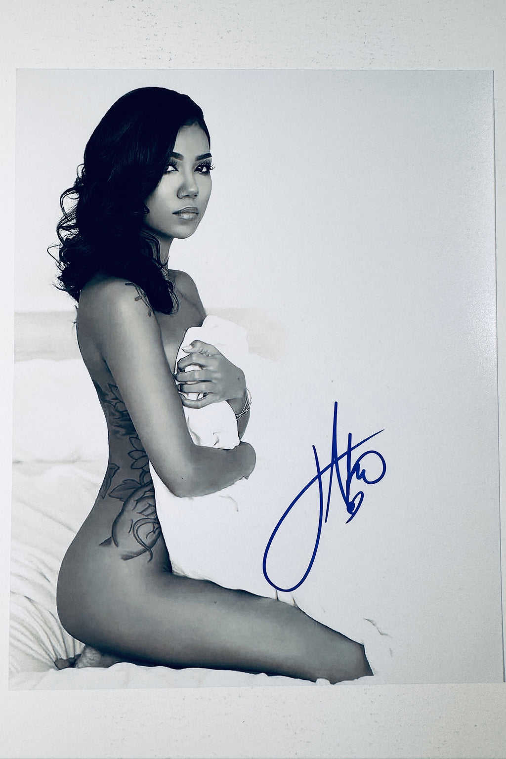 Signed full photo of Jhene Aiko posing kneeling on a bed while wrapped in a white sheet, featuring a blue autograph.