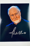 Signed 8x10 matte promotional portrait of legendary film composer John Williams wearing glasses and a dark shirt against a blue background, featuring a bold silver ink autograph.