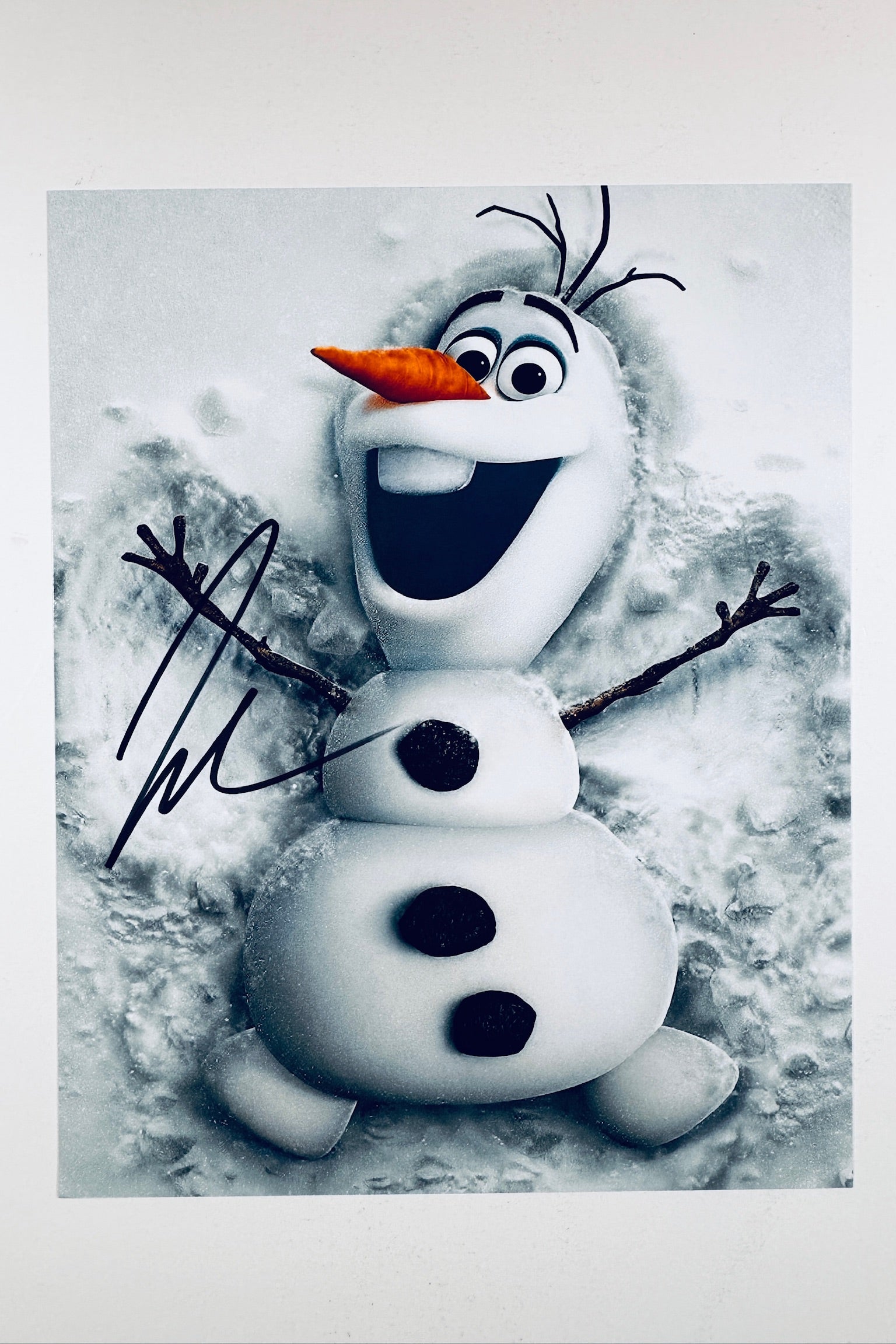 A promotional image of Olaf from Frozen smiling and lying in the snow, featuring a black autograph signed across the left side.