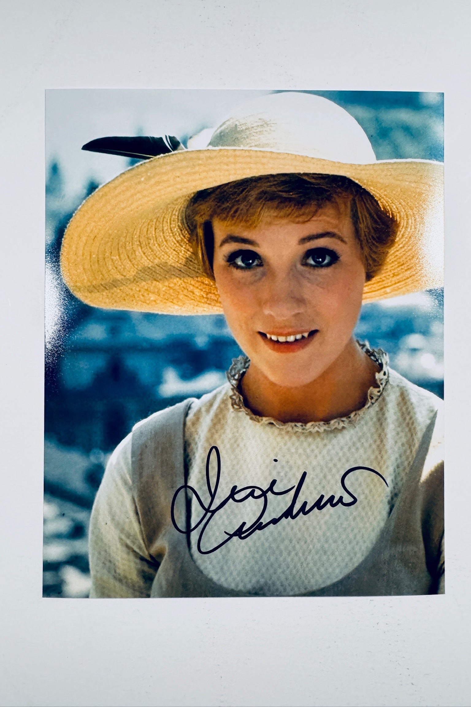 A close-up portrait of Julie Andrews wearing a wide-brimmed straw hat with a black feather and a light-colored dress in the classic movie The Sound of Music, featuring a dark marker signature across the lower center.