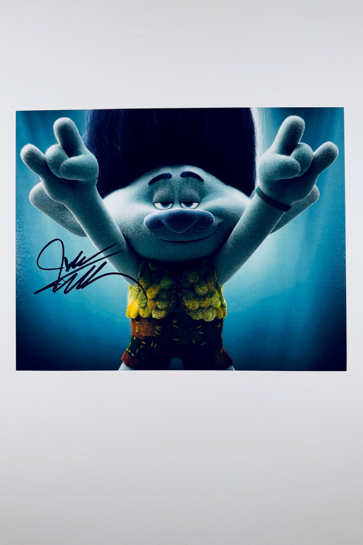 A still featuring Branch from Trolls holding his hands up making rock-on hand signs against a blue background, signed in black marker on the left side.