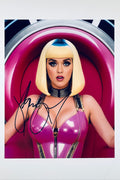 A portrait of Katy Perry wearing a pink latex outfit and a blonde bob wig with blue roots, sitting in a pink chair, featuring a signature in black marker on the left side.
