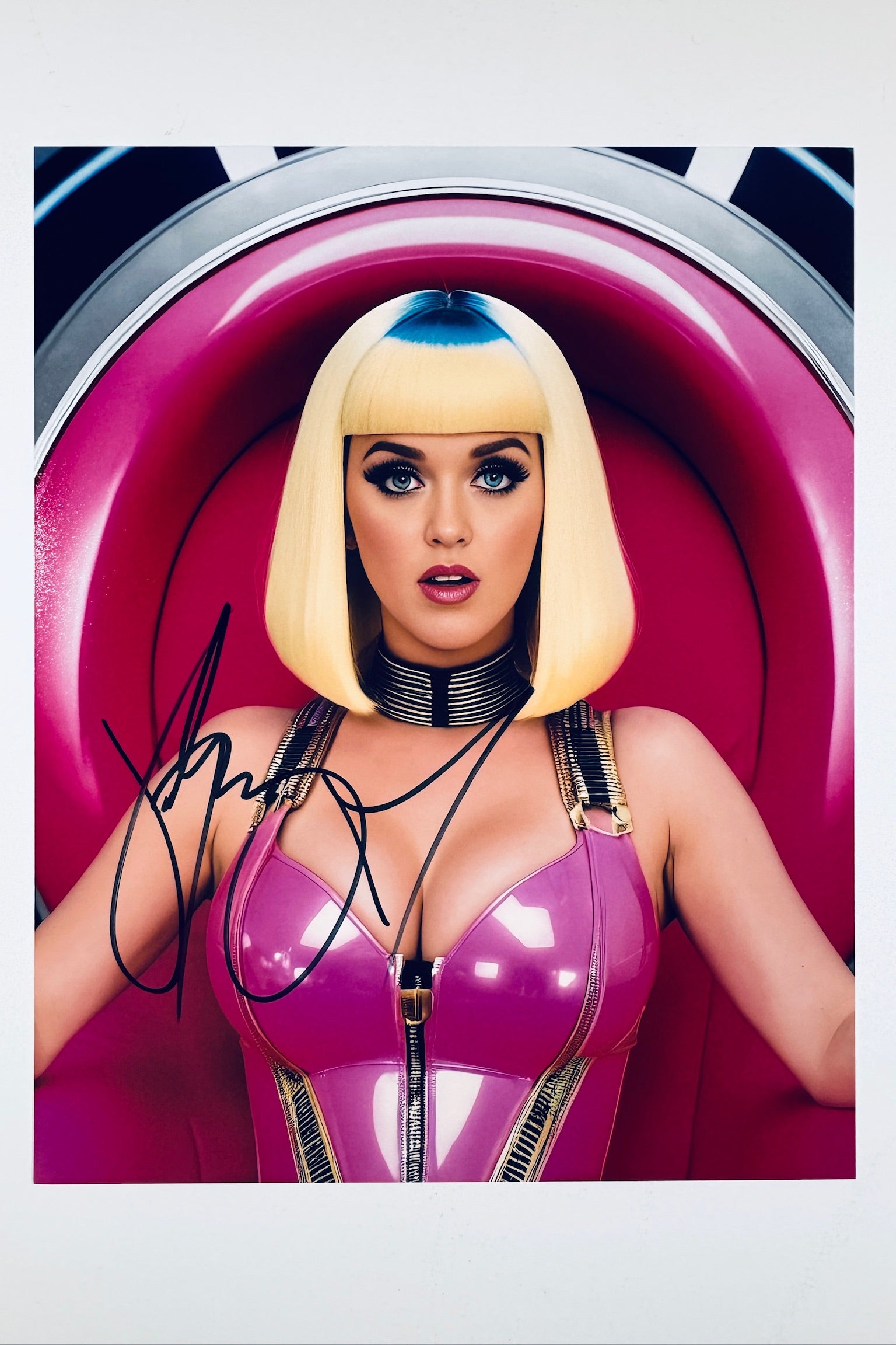 A portrait of Katy Perry wearing a pink latex outfit and a blonde bob wig with blue roots, sitting in a pink chair, featuring a signature in black marker on the left side.