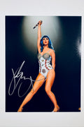 A full-length portrait of Katy Perry performing on stage with blue hair, wearing a sequined patriotic bodysuit and raising a microphone overhead against a dark background, featuring a silver marker signature across her lower left.