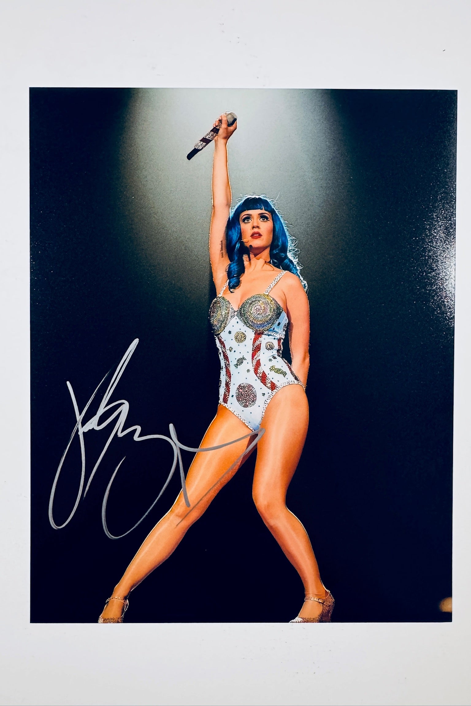 A full-length portrait of Katy Perry performing on stage with blue hair, wearing a sequined patriotic bodysuit and raising a microphone overhead against a dark background, featuring a silver marker signature across her lower left.