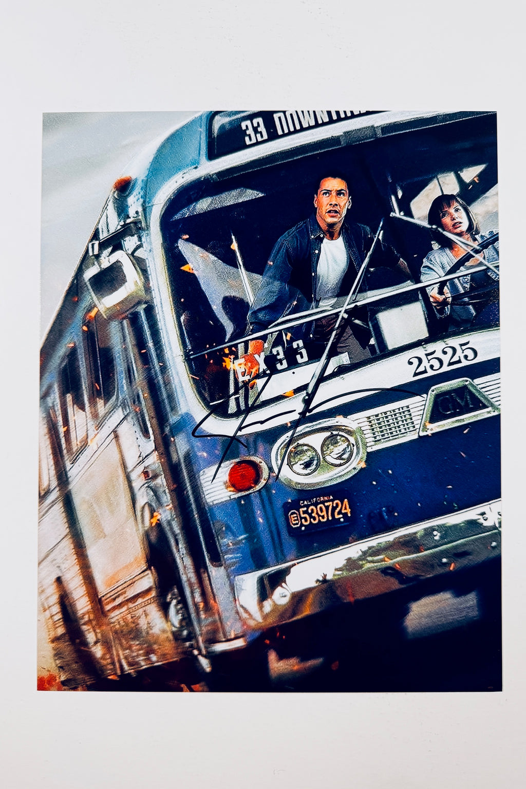 A promotional portrait of Keanu Reeves looking intensely forward from the driver's seat of a speeding blue bus with Sandra Bullock beside him, featuring a silver marker signature across the windshield.