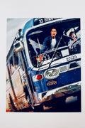 A promotional portrait of Keanu Reeves looking intensely forward from the driver's seat of a speeding blue bus with Sandra Bullock beside him, featuring a silver marker signature across the windshield.