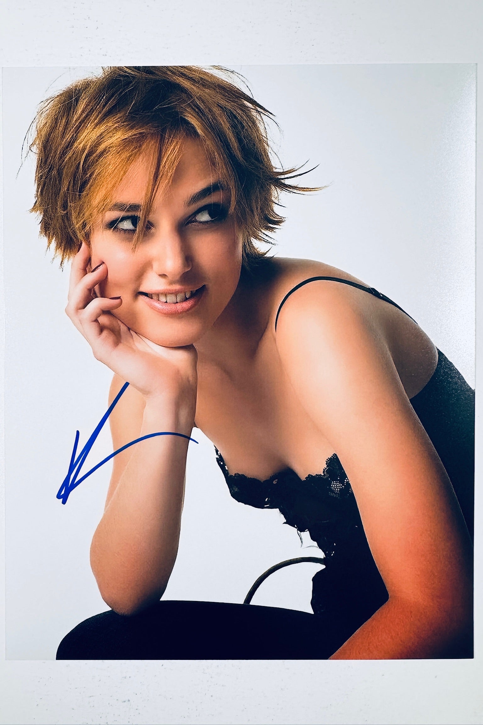 Signed 8x10 matte promotional photo of actress Keira Knightley with short brown hair resting her head in her hand while wearing a dark lacy top against a white background, featuring a bold blue ink autograph.