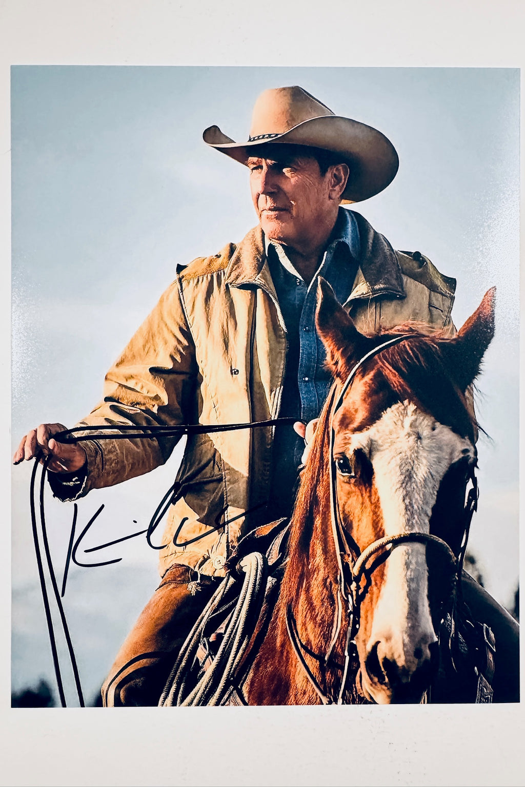 A portrait of Kevin Costner from the television series Yellowstone, wearing a cowboy hat, denim shirt, and tan jacket while riding a brown and white horse, featuring a dark marker signature on the lower left.