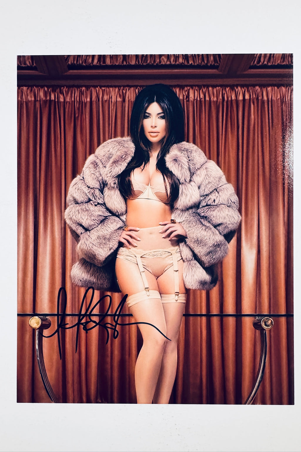 A portrait of Kim Kardashian wearing lingerie and a large fur coat standing in front of brown curtains with a glass table rail in the foreground, featuring a black marker signature across the lower left.