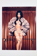 A portrait of Kim Kardashian wearing lingerie and a large fur coat standing in front of brown curtains with a glass table rail in the foreground, featuring a black marker signature across the lower left.