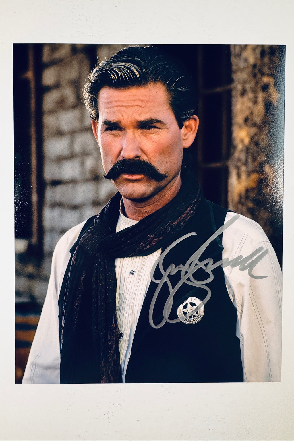 Signed 8x10 glossy photo of Kurt Russell as Wyatt Earp with a prominent mustache and Deputy Marshal badge from the movie Tombstone, featuring a silver ink autograph.