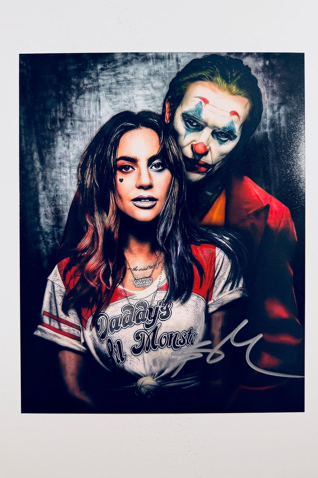 A portrait of Lady Gaga as Harley Quinn with a heart tattoo on her cheek and a Joker in clown makeup standing behind her, featuring a silver marker signature across the lower right.