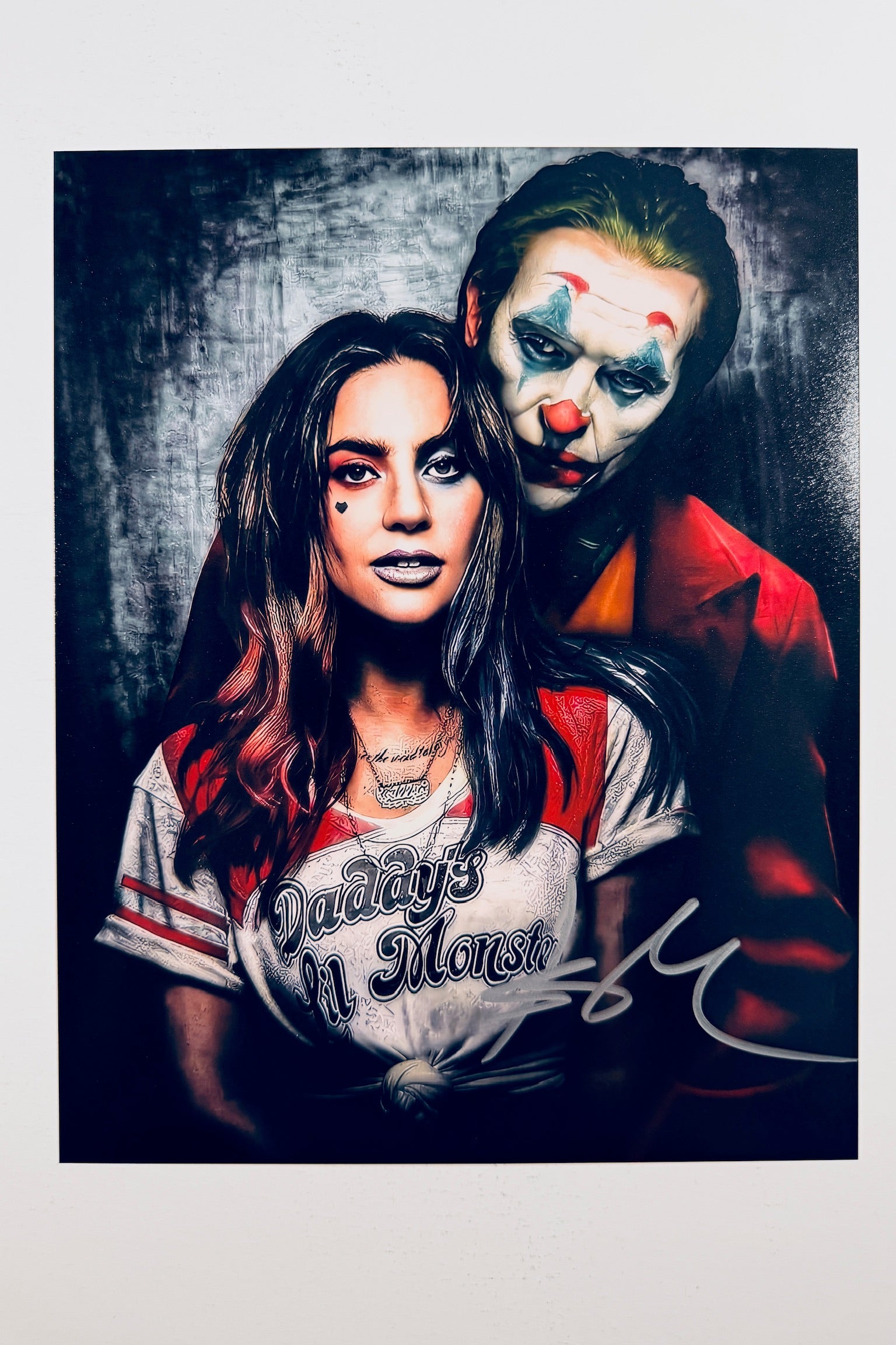 A portrait of Lady Gaga as Harley Quinn with a heart tattoo on her cheek and a Joker in clown makeup standing behind her, featuring a silver marker signature across the lower right.