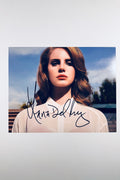 A portrait of Lana Del Rey with auburn hair wearing a white collared blouse against a blue sky, featuring a black marker signature across the front center.