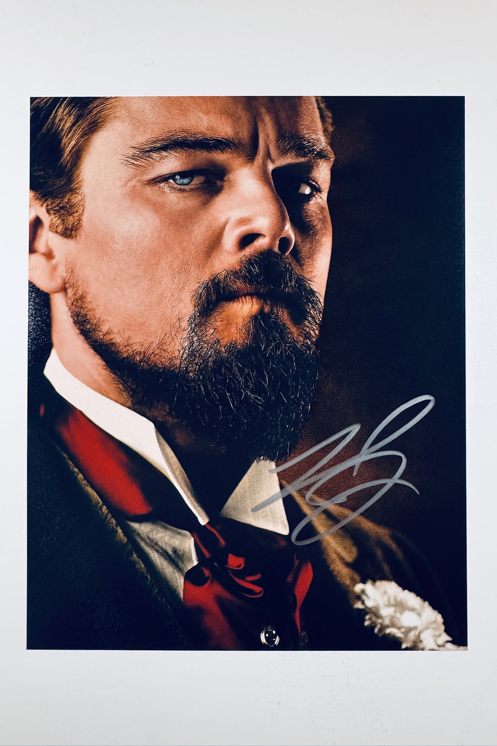 A close-up portrait of Leonardo DiCaprio as Calvin Candie from Django Unchained with a styled beard and mustache, wearing a dark suit with a red ascot and a white flower boutonniere, featuring a silver autograph in the lower right corner.