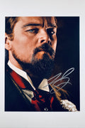 A close-up portrait of Leonardo DiCaprio as Calvin Candie from Django Unchained with a styled beard and mustache, wearing a dark suit with a red ascot and a white flower boutonniere, featuring a silver autograph in the lower right corner.