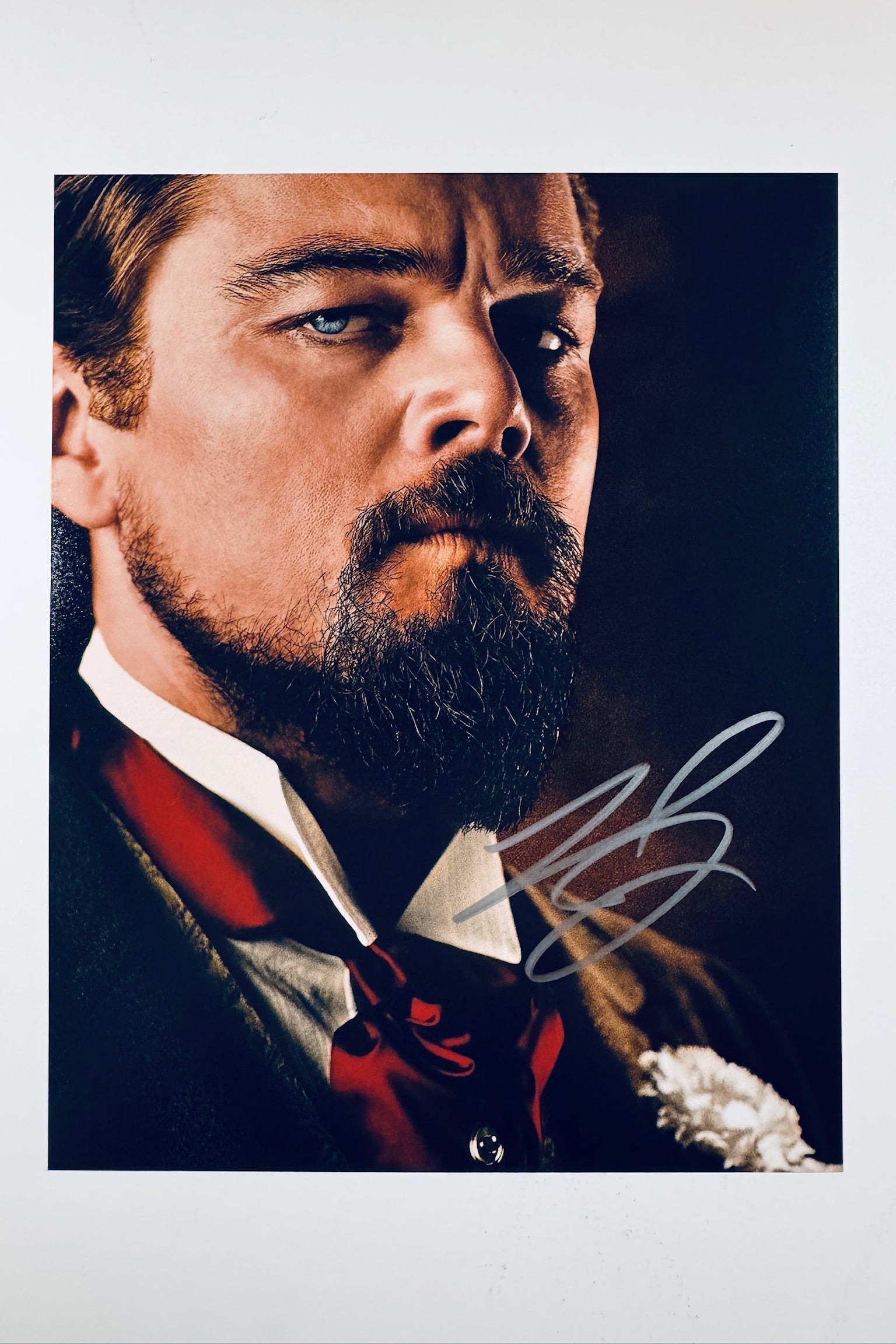 A close-up portrait of Leonardo DiCaprio as Calvin Candie from Django Unchained with a styled beard and mustache, wearing a dark suit with a red ascot and a white flower boutonniere, featuring a silver autograph in the lower right corner.