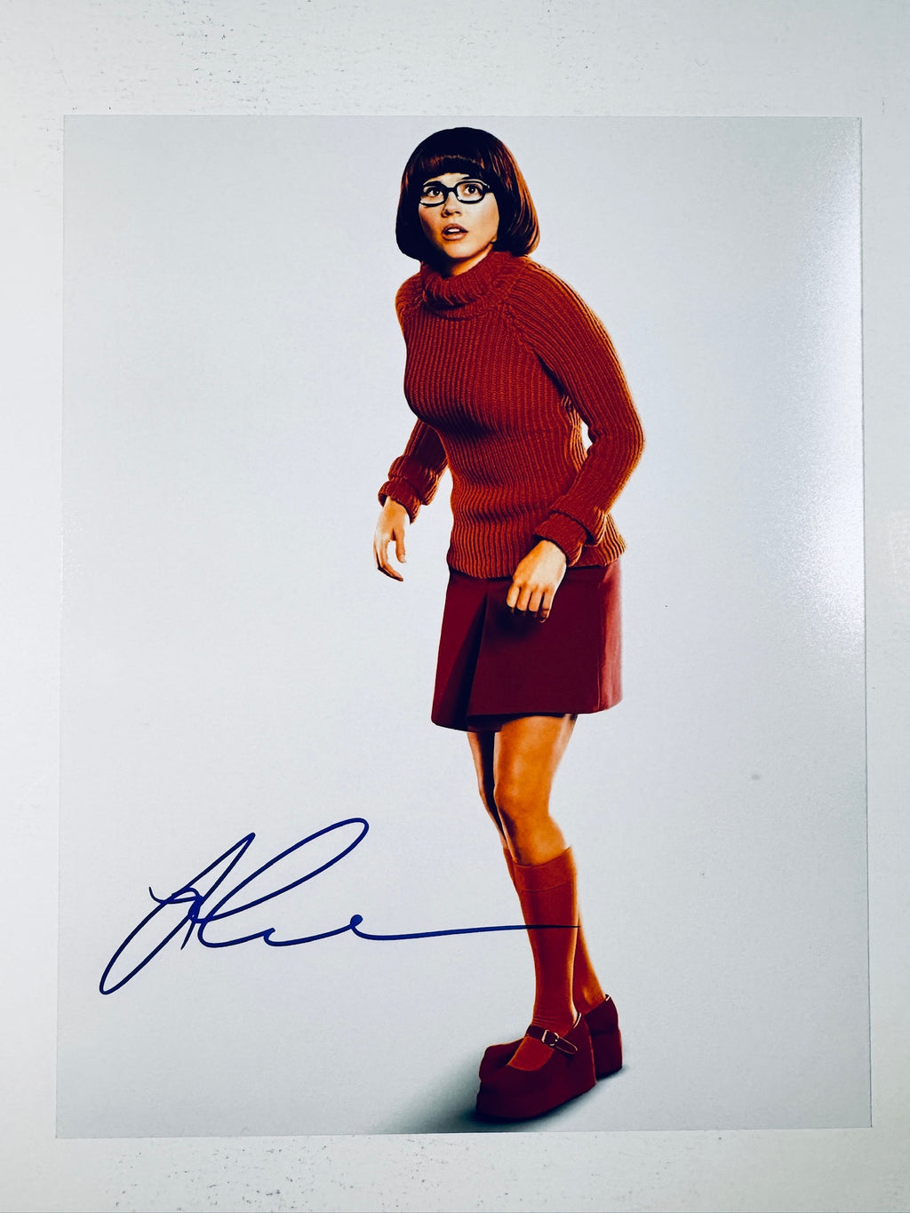 Signed 8x10 matte photo of Linda Cardellini as Velma from Scooby-Doo wearing her signature orange turtleneck, red skirt, and glasses, featuring a bold blue ink autograph across the lower-left corner.