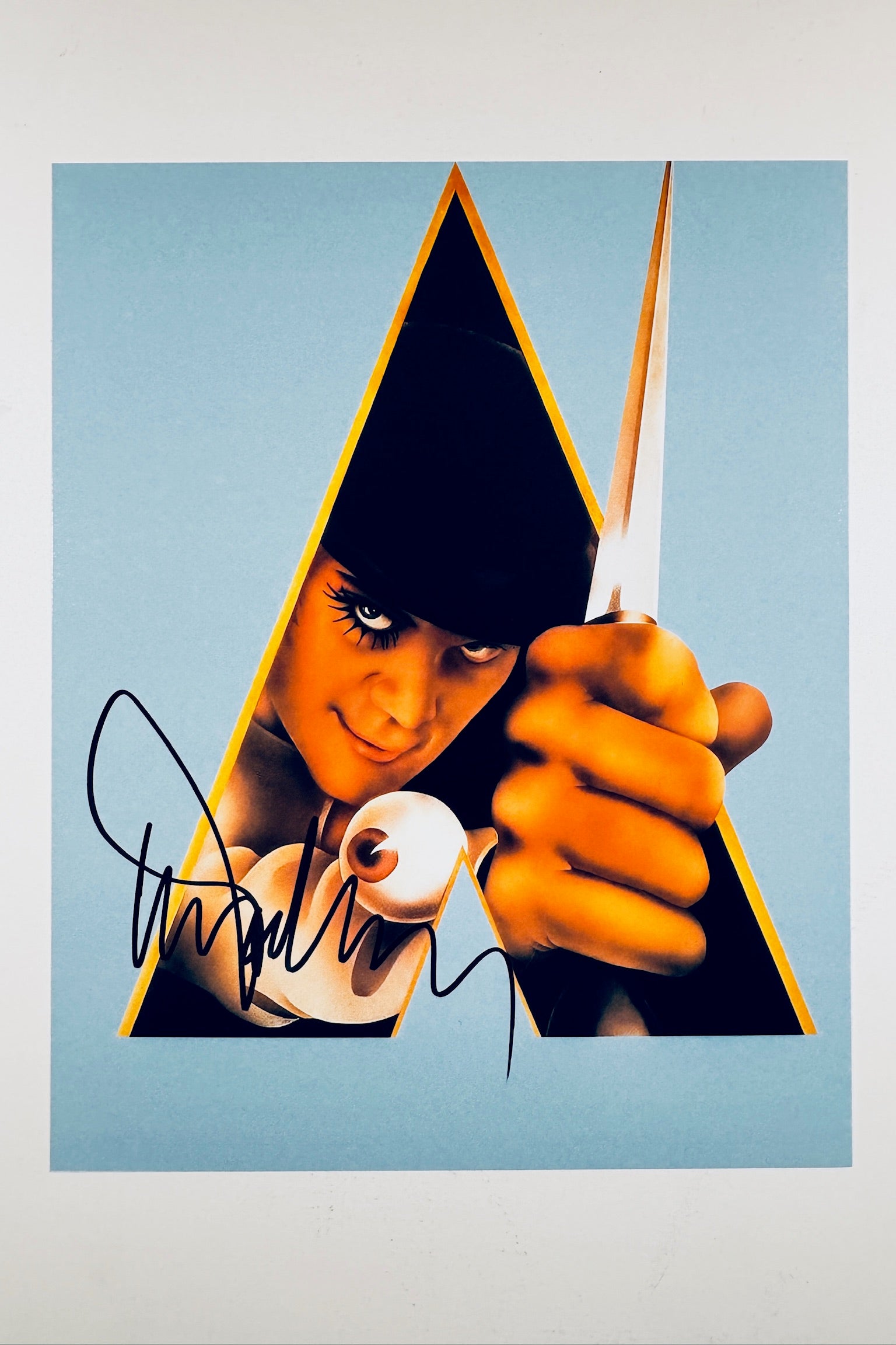 A portrait of Malcolm McDowell as Alex DeLarge from A Clockwork Orange, holding a knife and featuring a signature in black ink on the lower left.