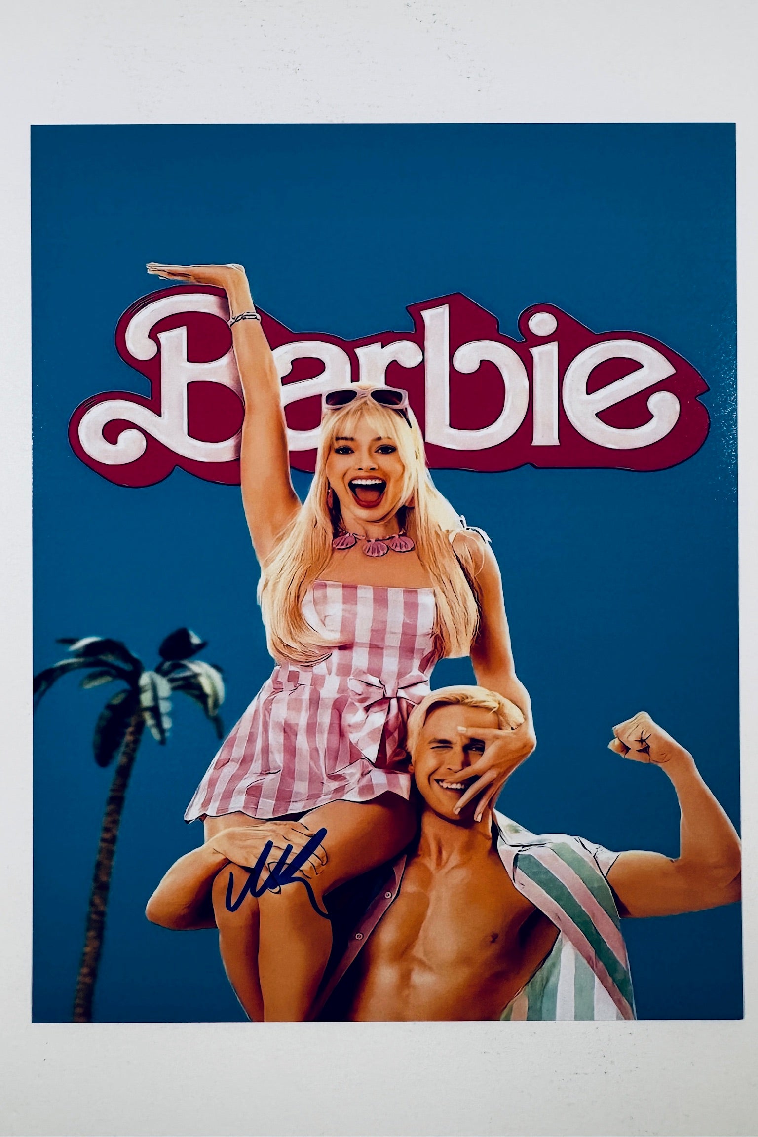 A promotional poster portrait for Barbie featuring Margot Robbie as Barbie in a pink gingham dress sitting atop Ryan Gosling as Ken with a blue background, featuring a blue marker signature across the lower left.