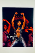 A performance portrait of Megan Thee Stallion with dark curly hair wearing a patterned, form-fitting stage outfit while flanked by two backup dancers on a dark stage, featuring a silver marker signature across the lower portion.