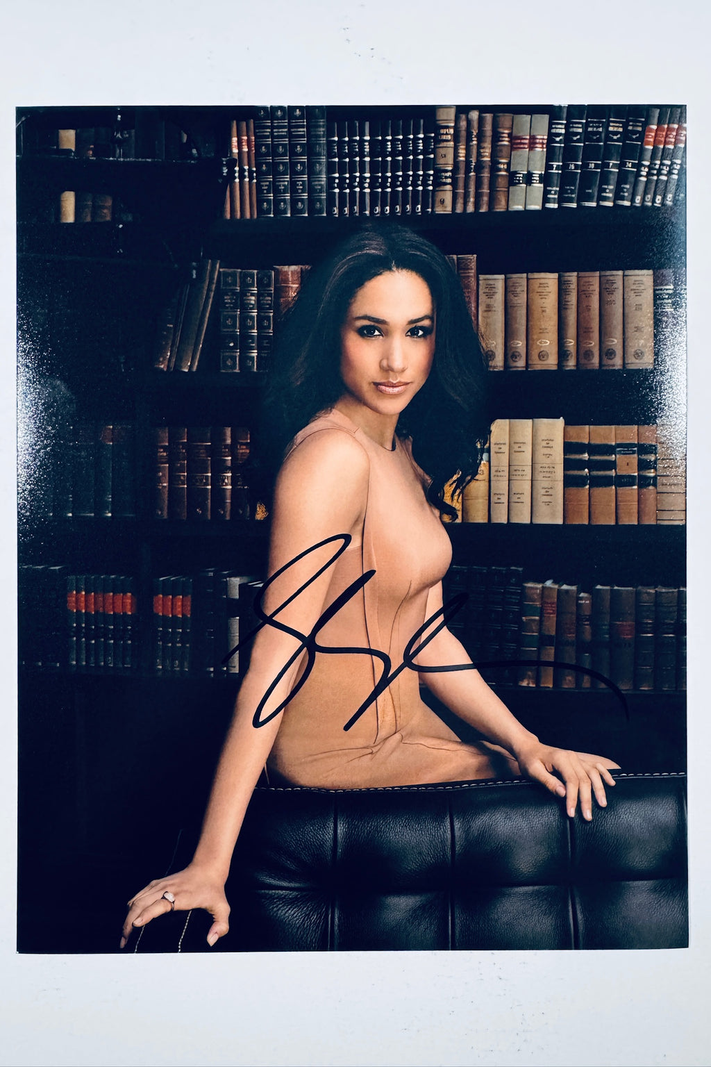A portrait of Meghan Markle wearing a beige sleeveless dress and seated on a black leather couch in front of a dark bookshelf filled with books, featuring a black marker signature across her lower lap.
