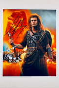 A promotional portrait of Mel Gibson as William Wallace in Braveheart wearing leather armor and a plaid kilt, holding a broadsword against an orange and fiery background, featuring a bold black autograph signed across the upper left area.