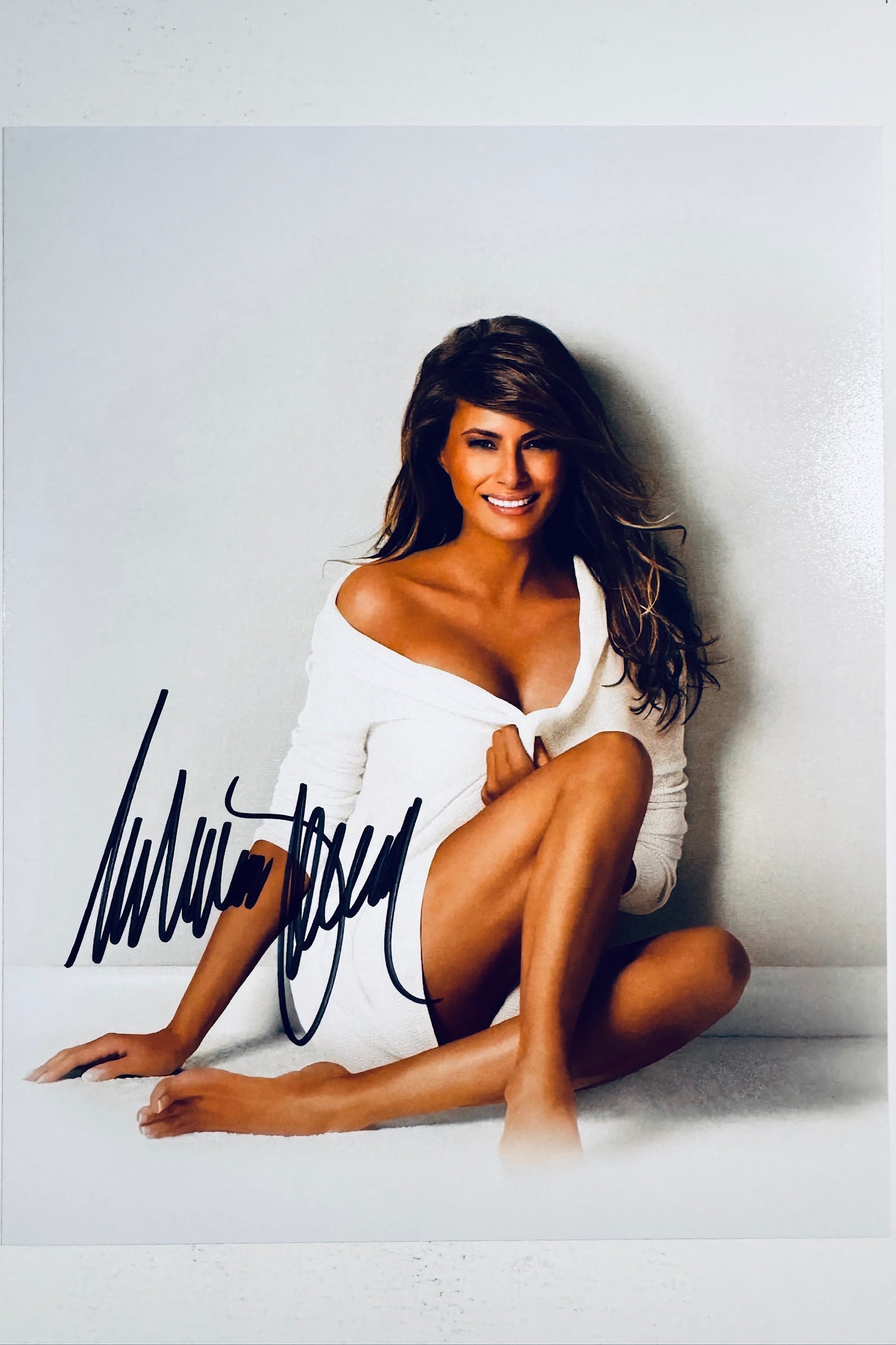 A portrait of Melania Trump sitting on a white floor against a plain wall, wearing a white off-the-shoulder top and smiling towards the camera, featuring a black marker signature on the lower left.