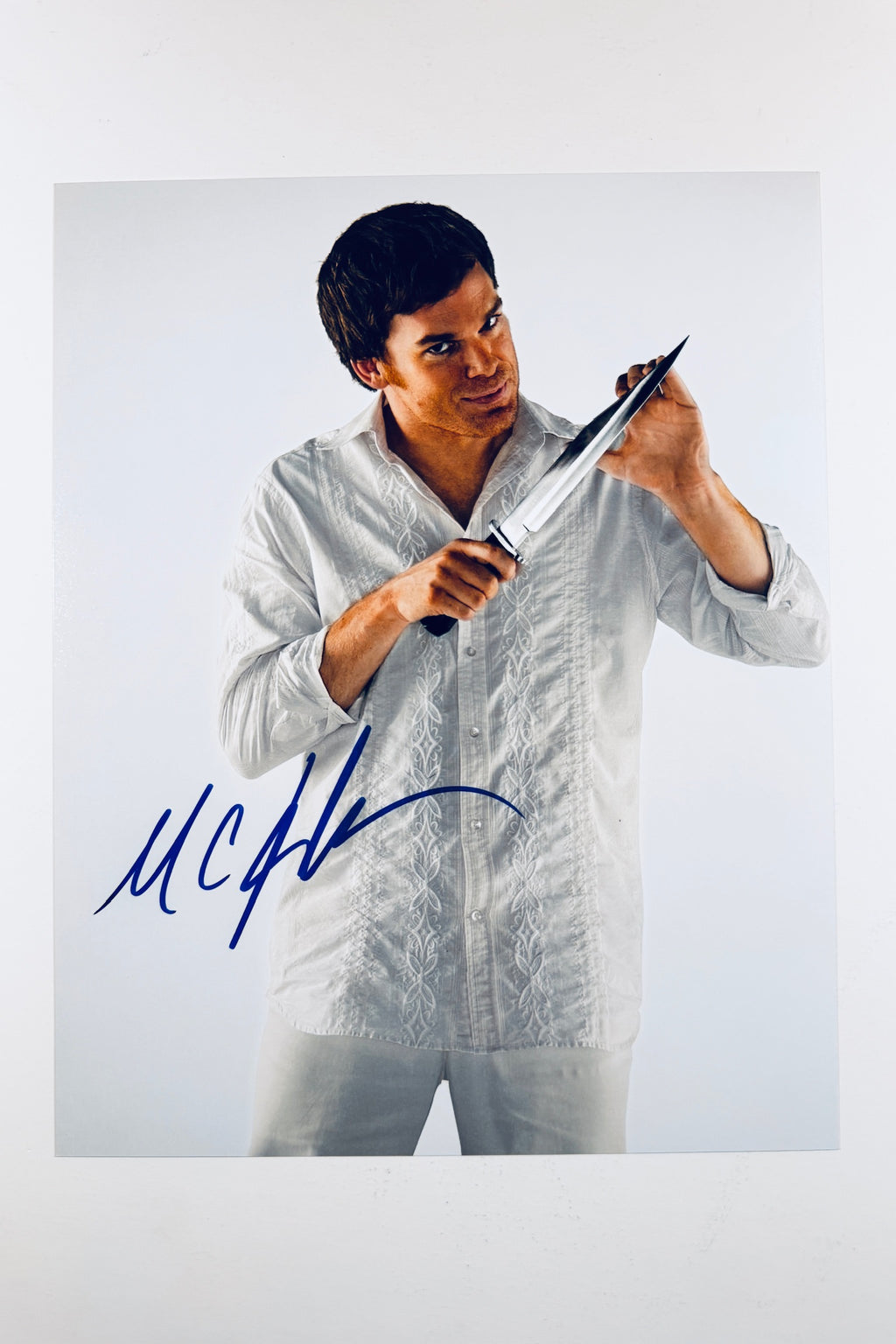 An autographed photo print of actor Michael Peña holding a large knife, wearing a white textured shirt against a light gray background, with a blue signature at the bottom left.