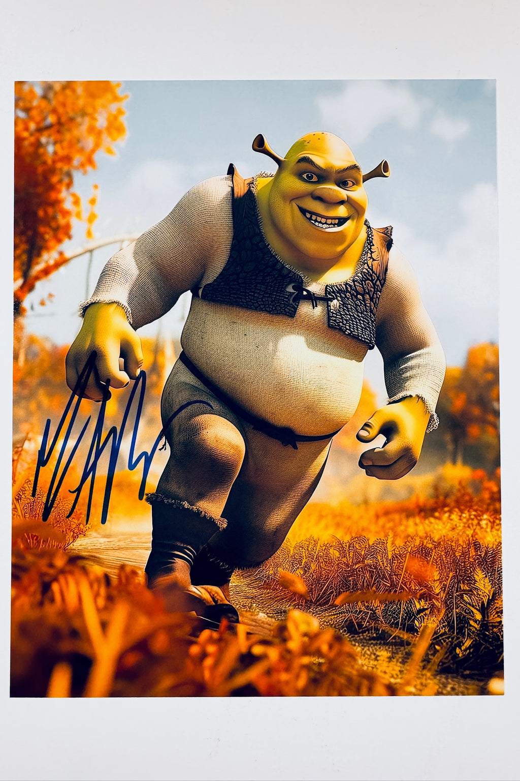 An autographed photo print of Shrek in his running pose from 'The Fairly OddParents', featuring a blue ink signature on the left side. The character is depicted in an outdoor autumn landscape with orange foliage under a blue sky.