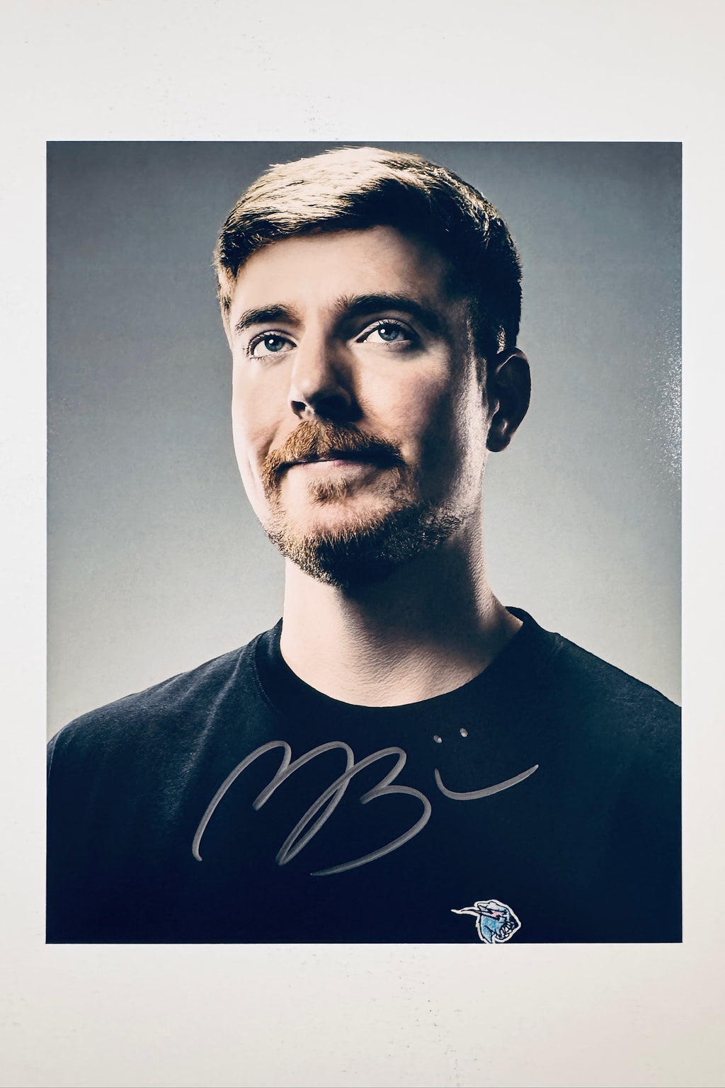 A close-up portrait of Mr. Beast with a slight smile wearing a black t-shirt against a grey background, featuring a bold black marker signature across the chest.