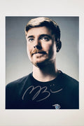 A close-up portrait of Mr. Beast with a slight smile wearing a black t-shirt against a grey background, featuring a bold black marker signature across the chest.