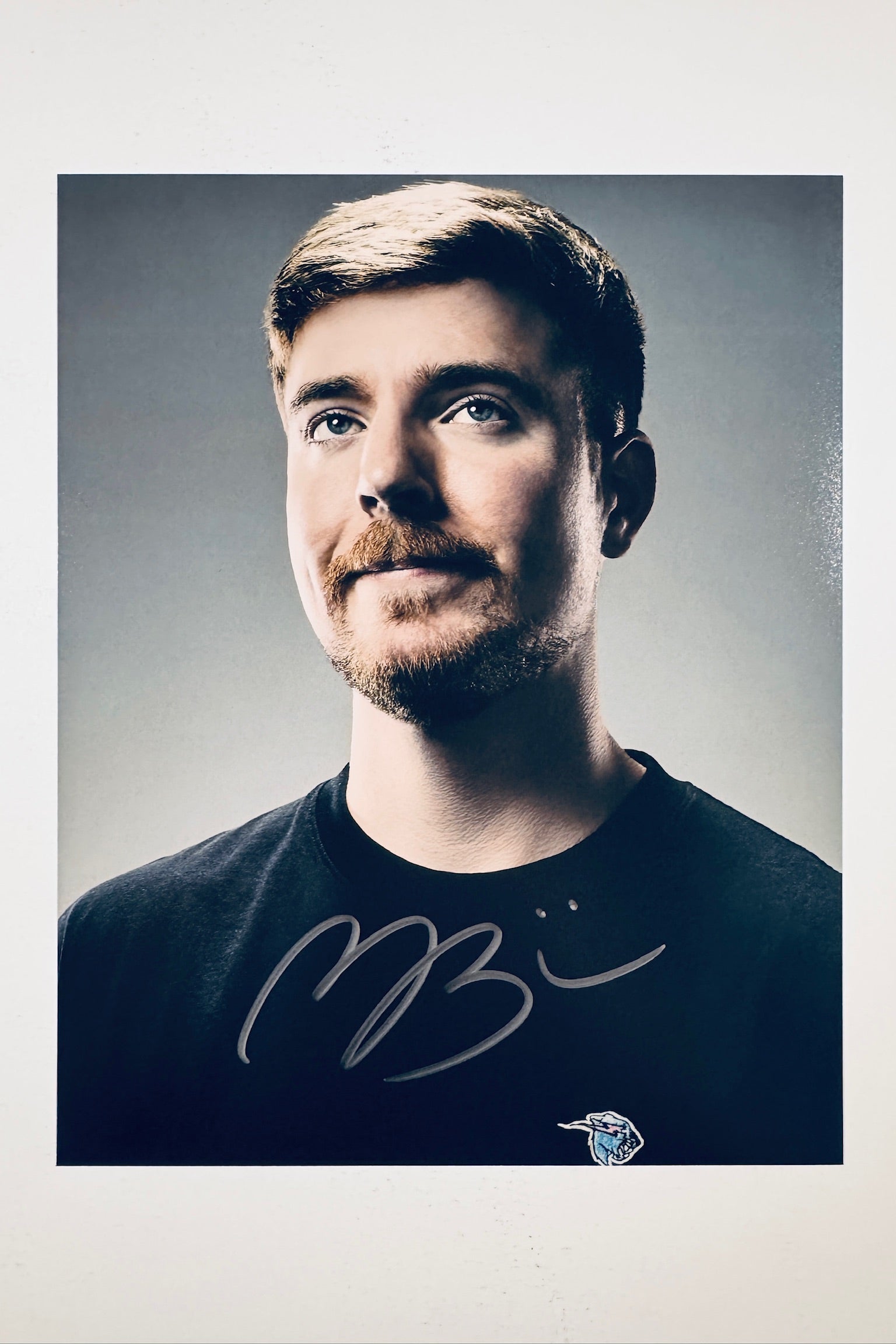 A close-up portrait of Mr. Beast with a slight smile wearing a black t-shirt against a grey background, featuring a bold black marker signature across the chest.