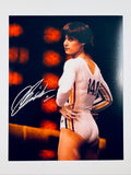 Signed 8x10 matte photo of Olympic gymnast Nadia Comaneci posing in a white and striped leotard with her hair short and dark, featuring a bold silver or white ink autograph across the left side.