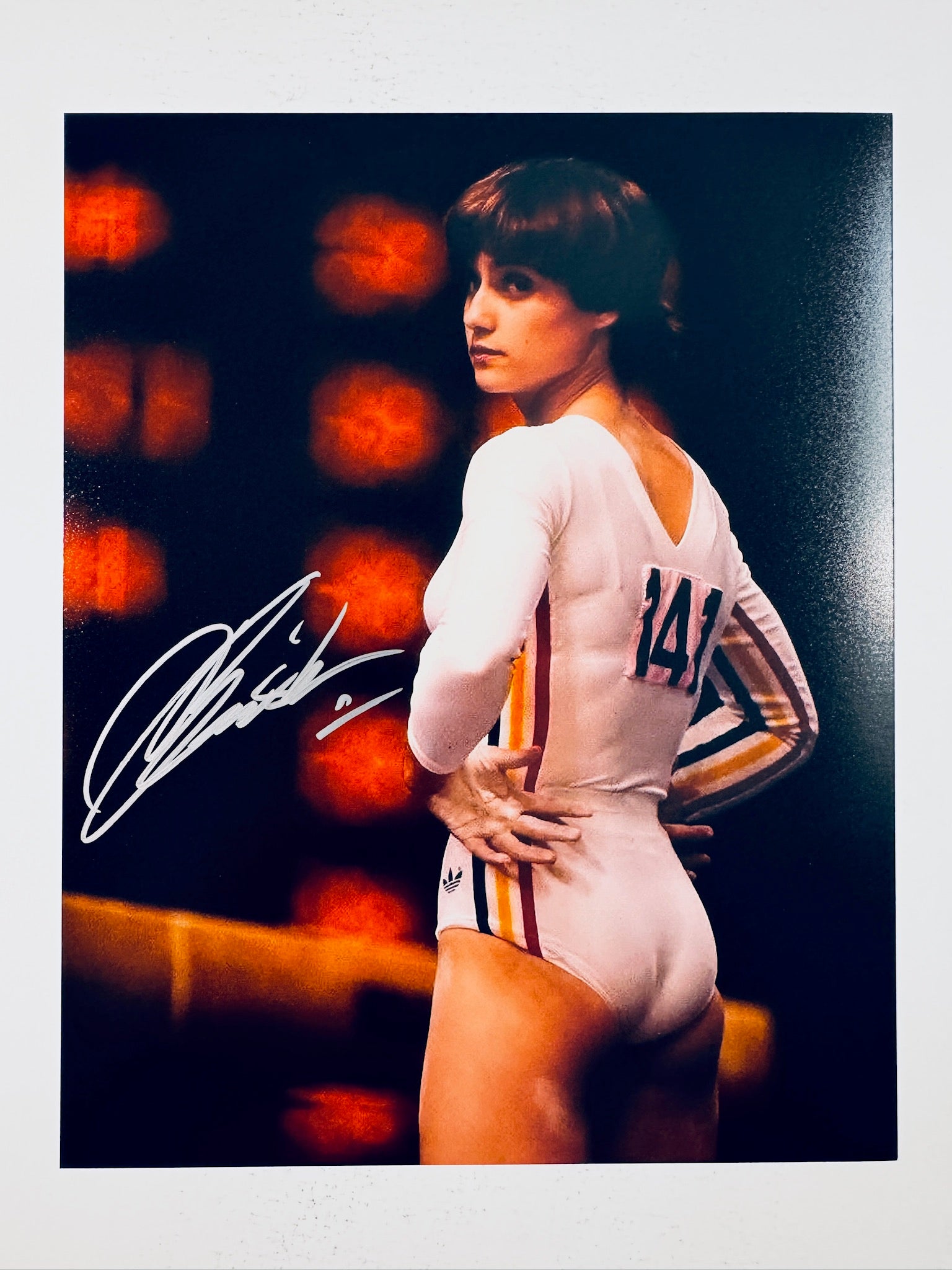Signed 8x10 matte photo of Olympic gymnast Nadia Comaneci posing in a white and striped leotard with her hair short and dark, featuring a bold silver or white ink autograph across the left side.
