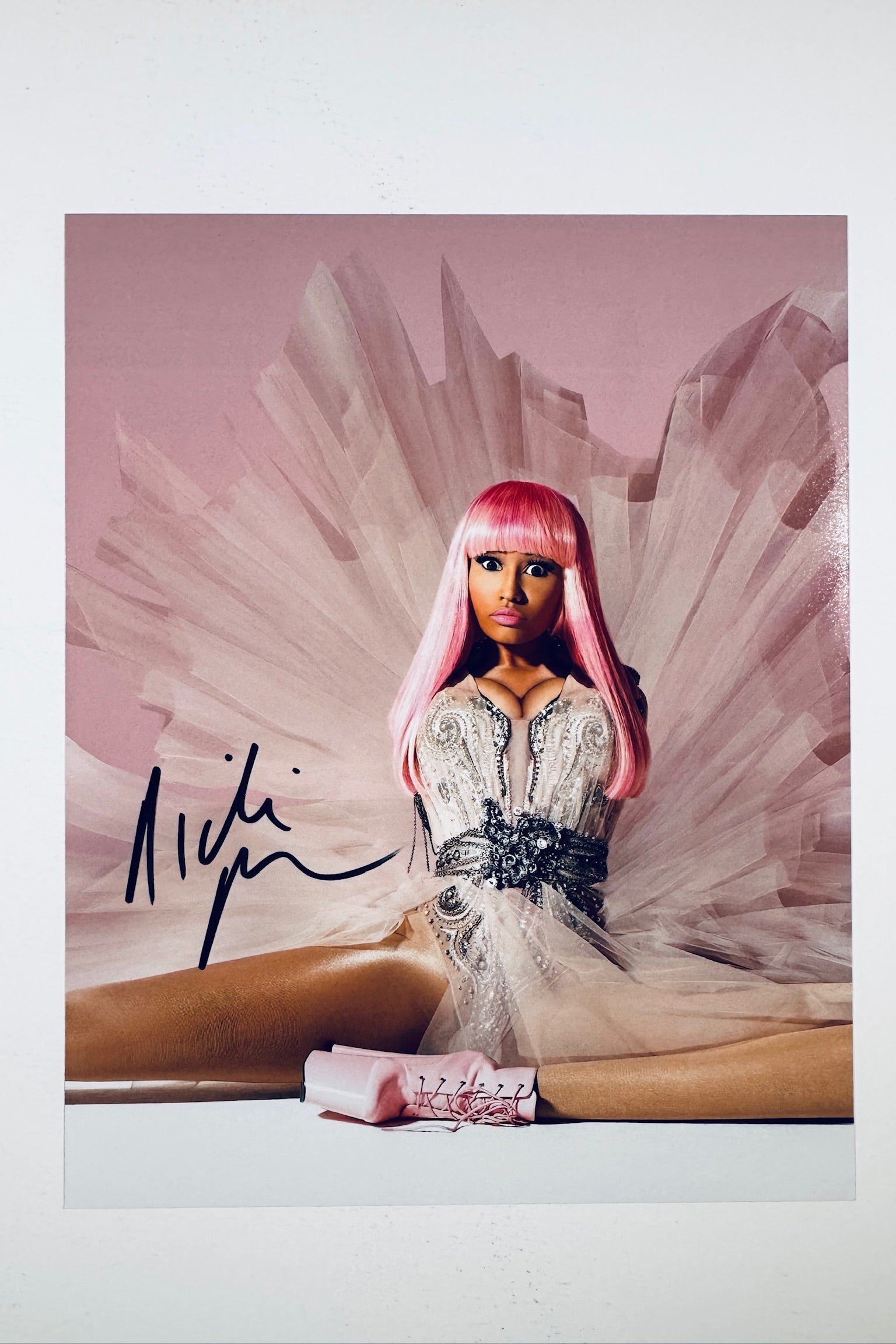 A portrait of Nicki Minaj with pink hair wearing an ornate beaded dress and pink platform boots, seated against a pale pink backdrop with an expansive ruffled tulle train, featuring a black marker signature across the left side.