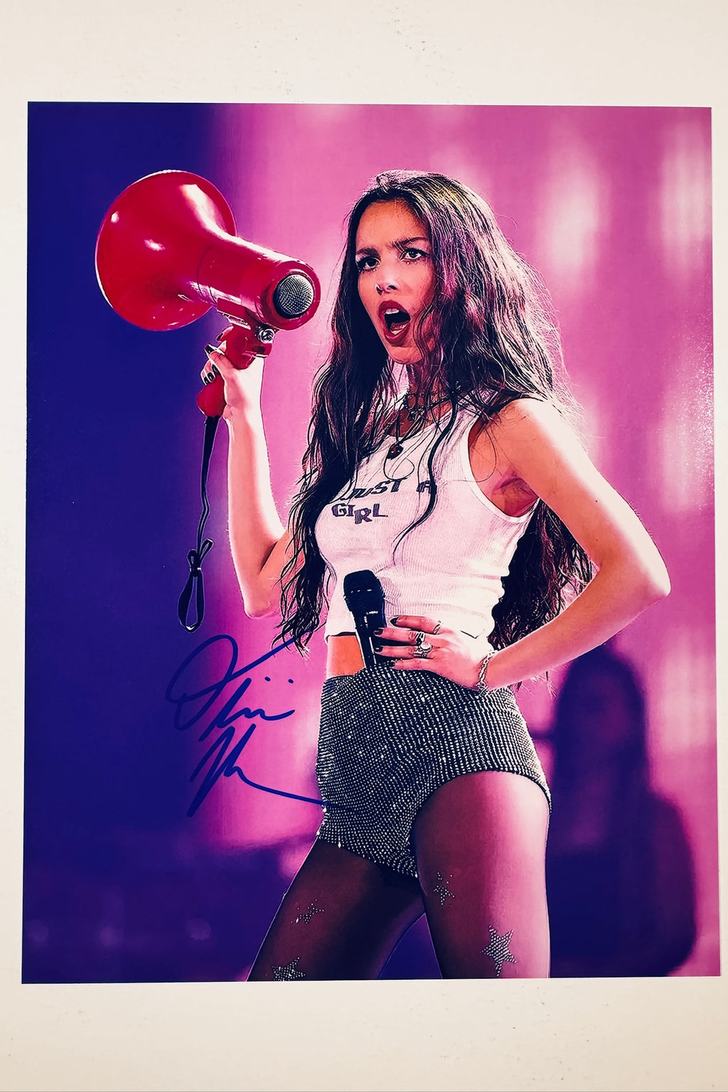 A portrait of Olivia Rodrigo singing live while holding a red megaphone and a microphone, wearing a white tank top and sparkly shorts against a pink background, featuring a blue marker signature across her lower left side.